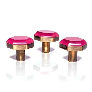 May include: Three decorative cabinet knobs with a geometric design. Each knob features a bright pink, faceted top, a gold-colored band, and a dark brown base. The knobs have a cylindrical, bronze-colored stem.