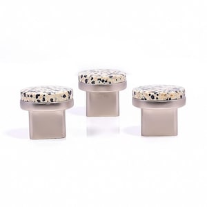 May include: Three cabinet knobs with a speckled, cream-coloured top and a silver-coloured base. The knobs have a circular top with black and brown spots. The base is rectangular and has a brushed metal finish. The knobs are displayed against a white background.