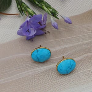 Pode incluir: Dois brincos de botão de pedra turquesa com molduras douradas. Os brincos são ovais e têm uma superfície facetada. Flores roxas e caules verdes no fundo.