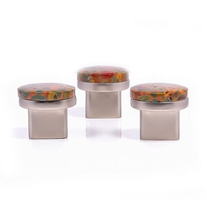 May include: Three cabinet knobs with a silver base and a colourful, speckled top. The tops feature a mix of red, orange, green, and yellow hues. The knobs are square-shaped and sit on a white background.