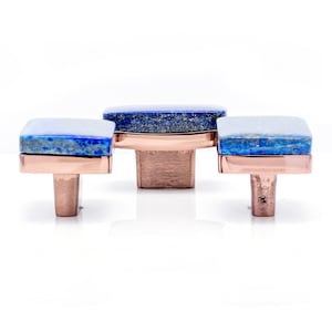 Könnte beinhalten: Drei Schrankknöpfe mit quadratischen Lapis-Lazuli-Aufsätzen und kupferfarbenen Metallsockeln. Die Knöpfe sind vor einem weißen Hintergrund in einer Reihe angeordnet. Der Lapis Lazuli hat blaue und weiße Marmorierungen.