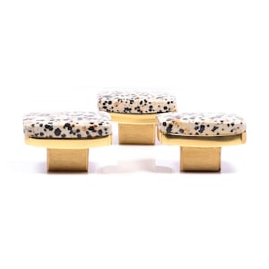 Könnte beinhalten: Drei Schrankknöpfe mit einer gesprenkelten, hellfarbigen Steinplatte und einem goldfarbenen Metallsockel. Die Knöpfe haben eine rechteckige Form und sind vor weißem Hintergrund in einer Reihe angeordnet. Diese dekorativen Knöpfe sind für Möbel geeignet.