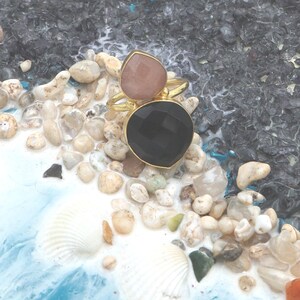 May include: A gold-toned ring featuring two gemstones: a teardrop-shaped pink stone and a large, round black stone. The ring is set against a backdrop of small pebbles, seashells, and a blue and white surface.