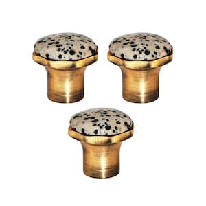 Op de afbeelding: Drie kastknoppen met een gespikkelde, lichtgekleurde stenen bovenkant en een goudkleurige, cilindrische basis. De knoppen hebben een klassiek ontwerp, geschikt voor meubels of kasten. De stenen bovenkant heeft een diameter van ongeveer 3 cm.