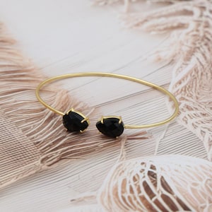 May include: A gold-tone open cuff bracelet with two black, teardrop-shaped gemstones. The bracelet is displayed on a light, textured surface. The gemstones are faceted, adding to the bracelet's visual appeal.