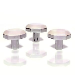 Pode incluir: Três puxadores decorativos para armários. Cada puxador apresenta um topo octogonal branco com uma borda de metal prateado e uma base cilíndrica. Os puxadores são exibidos sobre um fundo branco, com seus reflexos visíveis.