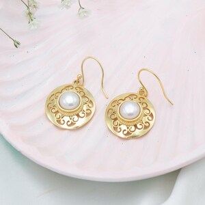 Könnte beinhalten: Ein Paar goldfarbene Ohrringe mit kreisförmigem Design. Jeder Ohrring hat eine zentrale weiße Perle, die von einem verzierten, durchbrochenen Muster umgeben ist. Die Ohrringe werden auf einer rosa Oberfläche mit kleinen weißen Blumen im Hintergrund präsentiert.
