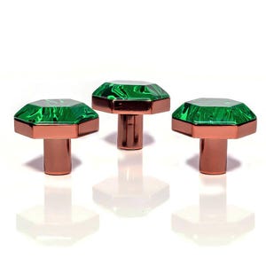 May include: Three cabinet knobs with a decorative design. Each knob has a faceted, emerald-green gem-like top and a copper-coloured base and stem. The knobs are set against a white background, with their reflections visible.