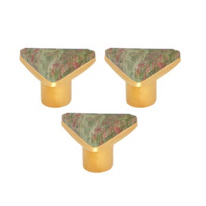 May include: Three cabinet knobs with a triangular top made of green and red speckled stone. The knobs have a gold-coloured metal base and trim, creating a decorative accent for furniture.