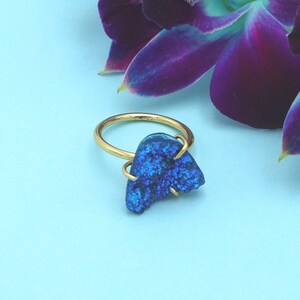 Op de afbeelding: Een goudkleurige ring met een levendige blauwe, onregelmatig gevormde steen. De ring staat tegen een lichtblauwe achtergrond, met een paarse orchidee in de rechterbovenhoek. De steen heeft een textuur, kristallijn uiterlijk.