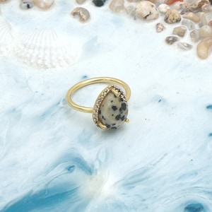 May include: A gold ring with a teardrop-shaped stone speckled with black spots. The stone is framed by small, clear gems. The ring sits on a blue and white surface, with seashells and pebbles in the background.