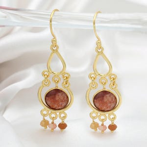 Pode incluir: Um par de brincos dourados de lustre. Cada brinco apresenta uma parte superior em forma de gota, um centro circular com uma pedra castanho-avermelhada e três contas penduradas em tons de amarelo, rosa e castanho.