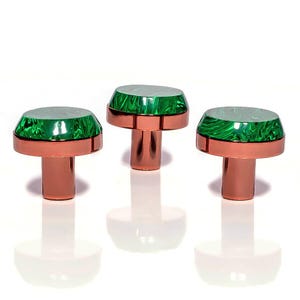 May include: Three cabinet knobs with a copper-coloured base and stem, each topped with a green, marbled, oval accent. The knobs are arranged in a row against a white background, with their reflections visible below. The knobs are likely for furniture.