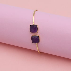 May include: A gold-colored bracelet with two square, faceted, deep purple gemstones. The bracelet has a simple, open design, with the gemstones set in gold-colored bezels. The background is a solid pink color.