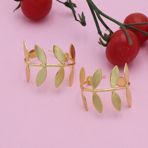 May include: A pair of gold-coloured leaf-shaped earrings. Each earring has a curved bar with several leaf-shaped elements attached. The earrings are set against a soft pink background, with red cherry tomatoes and green stems in the background.