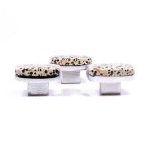 May include: Three square cabinet knobs with a speckled, cream-coloured stone top and a silver-coloured base. The knobs are arranged in a row against a white background. These decorative knobs are ideal for furniture upgrades.