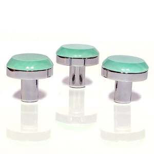 May include: Three cabinet knobs with a mushroom design. Each knob has a mint green top and a silver-coloured base and stem. The knobs are displayed on a white surface, reflecting the light.