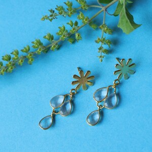 Könnte beinhalten: Ein Paar goldfarbene Ohrringe mit floralem Design und drei tropfenförmigen, hellblauen Edelsteinen. Die Ohrringe werden auf einem hellblauen Hintergrund präsentiert, mit einem Zweig grünen Laubes in der oberen linken Ecke.