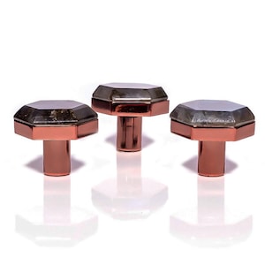 Könnte beinhalten: Drei dekorative Schrankknöpfe mit geometrischem Design. Jeder Knopf hat einen kupferfarbenen Sockel und eine facettierte, rauchgraue Oberseite. Die Knöpfe sind auf weißem Hintergrund angeordnet und zeigen ihre reflektierenden Oberflächen und ihr elegantes Design.