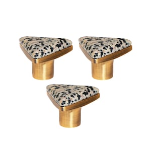 Op de afbeelding: Drie kastknoppen met een driehoekige bovenkant met een gespikkeld patroon in zwart, beige en crème. De knoppen hebben een messingkleurige basis en afwerking en staan tegen een witte achtergrond. Deze decoratieve knoppen zijn geschikt voor meubels.