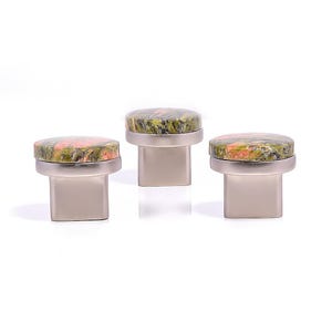 May include: Three cabinet knobs with a natural stone appearance. The knobs have a round top with a speckled pattern of green, pink, and orange hues. The base is a rectangular shape with a brushed metal finish.