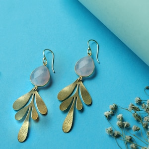 Puede incluir: Un par de pendientes dorados con una piedra preciosa azul claro y detalles en forma de hojas. Los pendientes tienen cierre de gancho y están sobre un fondo azul. Un artículo de joyería.