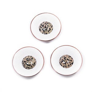 Pode incluir: Três bases brancas com um design de círculos concêntricos e uma borda cor de cobre. Cada base apresenta uma pedra central salpicada, criando um sotaque único e decorativo. Ideal para proteger superfícies.