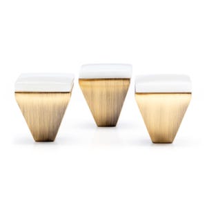 May include: Three decorative cabinet knobs with a geometric design. Each knob features a white, square top and a brushed brass-colored, inverted trapezoid base. The knobs are set against a white background.