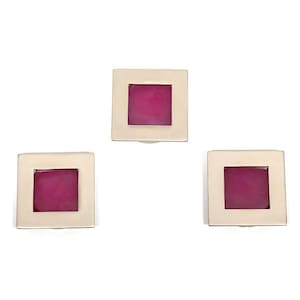 May include: Three square-shaped earrings with a silver-colored frame and a magenta-colored center. The earrings are arranged in a triangular pattern against a white background. The earrings are likely for pierced ears.