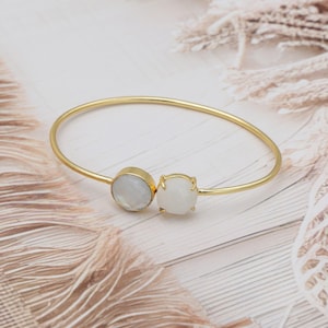 May include: A gold-toned bangle bracelet featuring two white gemstone accents. One is round, the other is a faceted square. The bracelet has a delicate, minimalist design, perfect for everyday wear.