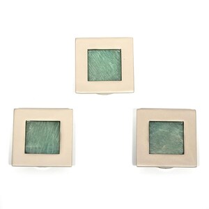 Könnte beinhalten: Drei quadratische Schrankknöpfe mit einem hellbeigen Rahmen und einem quadratischen grün-blauen Glas in der Mitte. Die Knöpfe sind vor einem weißen Hintergrund angeordnet.
