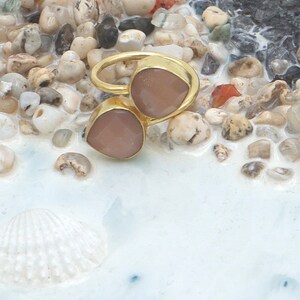 Può includere: Un anello dorato con due pietre preziose rosa chiaro a forma di goccia. L'anello è posto su uno sfondo di piccoli ciottoli colorati e una superficie bianca ornata di conchiglie, creando un'estetica da spiaggia.