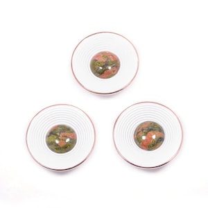 Puede incluir: Tres objetos decorativos redondos. Cada uno tiene un anillo exterior blanco y estriado con un borde color cobre. El centro de cada objeto presenta una piedra moteada multicolor en tonos verdes, rojos y marrones.