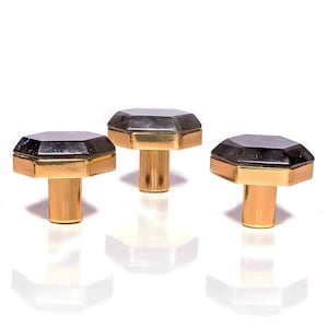 May include: Three cabinet knobs with a dark grey, faceted top and a gold-coloured base and stem. The knobs are displayed on a white background. Each knob is a decorative item.