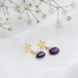 Könnte beinhalten: Goldfarbene sternförmige Ohrringe mit einem ovalen, tiefvioletten Stein. Die Ohrringe sind vor einem weißen Hintergrund mit zarten weißen Blumen platziert.