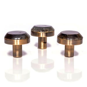 Peut inclure: Trois boutons de meuble en laiton avec un design en forme de champignon. Chaque bouton a un dessus sombre et une base en laiton. Les boutons sont disposés en rangée, projetant de doux reflets sur la surface blanche.