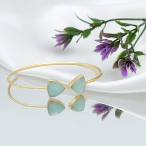 May include: A gold-coloured bangle bracelet featuring two light blue triangular stones. The bracelet is set against a white background with purple flowers and green leaves.