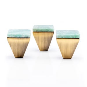 Może przedstawiać: Trzy dekoracyjne gałki do szafek z kwadratowym, jasnozielonym kamiennym blatem i mosiężną, odwróconą trapezową podstawą. Gałki są ustawione na białym tle, co podkreśla ich unikalny design i kontrast kolorystyczny. Wymiary w centymetrach.