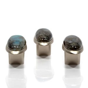 Può includere: Tre pomelli per mobili con top ovali in pietra preziosa iridescente nei toni del grigio, blu e verde. I pomelli hanno una base in metallo spazzolato e un design moderno. I pomelli sono esposti su sfondo bianco.