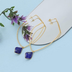 May include: A pair of gold-colored earrings with a geometric design. Each earring features a large, dark blue, gemstone pendant. The earrings are displayed on a reflective surface with purple flowers in the background. The earrings have a post-back closure.