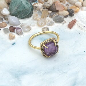 May include: A gold ring featuring a square, purple gemstone. The gemstone is framed by a square of smaller, dark stones. The ring is set against a light blue and white background.