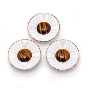 Puede incluir: Tres objetos redondos decorativos con un anillo exterior blanco y estriado y un borde cobrizo. Cada uno tiene un centro de piedra ojo de tigre marrón y dorado pulido. Los objetos están dispuestos en un patrón triangular sobre un fondo blanco.