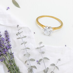 May include: A gold engagement ring with a large, clear gemstone. The ring is set against a white background with sprigs of lavender and eucalyptus leaves. The ring's band is a simple, elegant design.