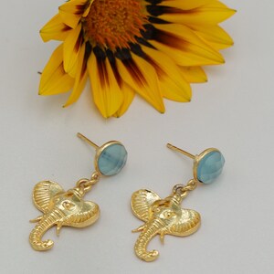 May include: A pair of gold-colored elephant earrings with light blue gemstone studs. The earrings are detailed with textured elephant heads hanging below the studs. A yellow flower with a dark center is in the background.