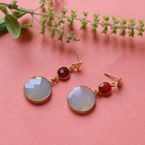 Może przedstawiać: Para złotych kolczyków o geometrycznym wzorze. Każdy kolczyk ma duży, okrągły, fasetowany, blado-niebieski kamień szlachetny, mniejszy czerwony kamień szlachetny i złoty trójkątny słupek. Kolczyki są prezentowane na różowej powierzchni.