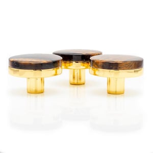May include: Three cabinet knobs with a decorative design. Each knob has a round top made of tiger-eye stone, set on a gold-coloured base and stem. The knobs are displayed on a white surface, highlighting their colours and design.