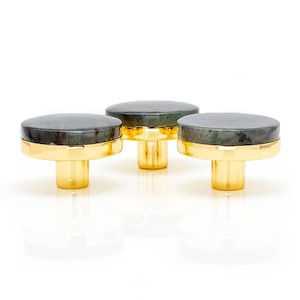 Könnte beinhalten: Drei Schrankknöpfe mit goldfarbenem Sockel und dunkelgrauer, kreisförmiger Oberseite. Die Knöpfe haben ein modernes Design und sind vor einem weißen Hintergrund platziert. Die Knöpfe sind wahrscheinlich aus Metall und Stein.