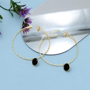 May include: A pair of gold-colored hoop earrings with a hammered texture. Each earring features a black, oval-shaped stone. The earrings are displayed on a light blue surface with a reflective background and purple flowers.