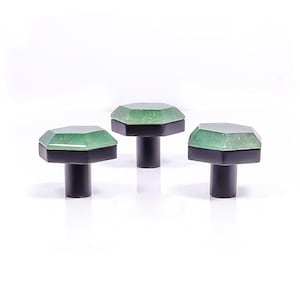 Può includere: Tre pomelli decorativi per mobili. Ogni pomello presenta una parte superiore sfaccettata in pietra preziosa verde chiaro, una base nera e uno stelo nero. I pomelli sono disposti in fila su sfondo bianco.