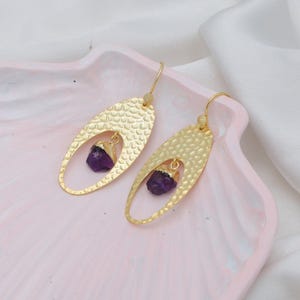 May include: Gold-tone earrings with an oval, textured design. Each earring has a raw amethyst gemstone suspended within the oval shape. The earrings have a hook fastening and a small decorative detail near the top.